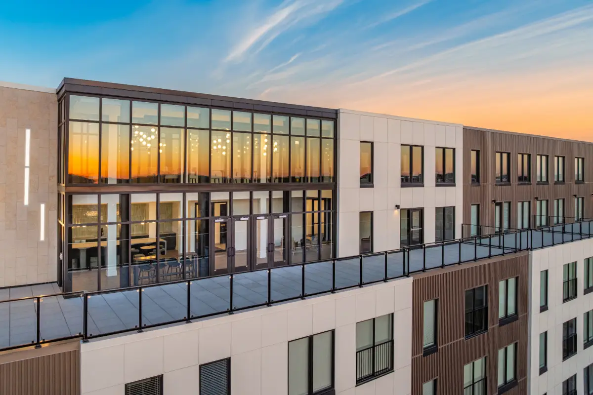 Modern building with large glass windows reflecting a vibrant sunset sky. Upper-level terrace with glass railing, creating a serene and luxurious atmosphere.