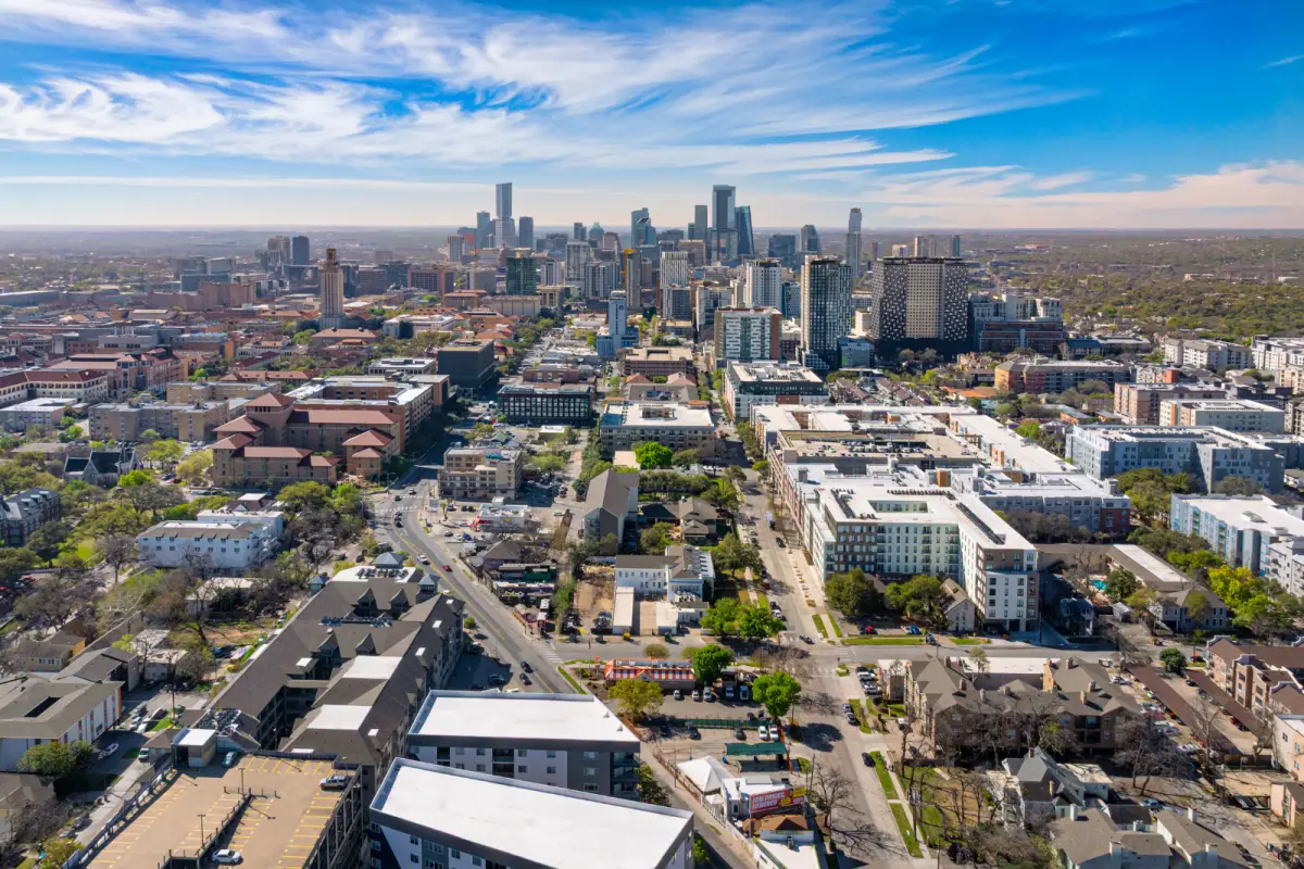 Aerial view of a cityscape with tall modern skyscrapers in the background under a blue sky. Foreground shows residential and commercial buildings.