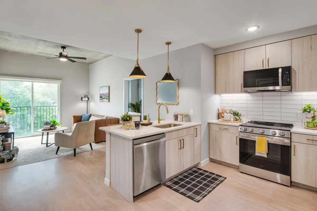 Modern kitchen and living room with light wood cabinets, black appliances, pendant lights, and neutral tones. Sunlit living area with cozy seating.
