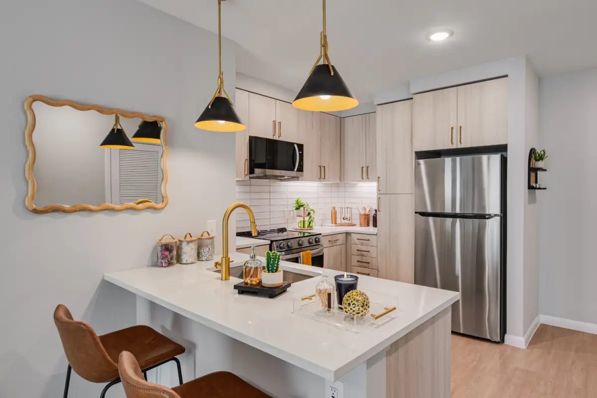 Modern kitchen with wooden cabinetry, stainless steel appliances, and a white island. Pendant lights hang above brown chairs. Reflective mirror adds depth.