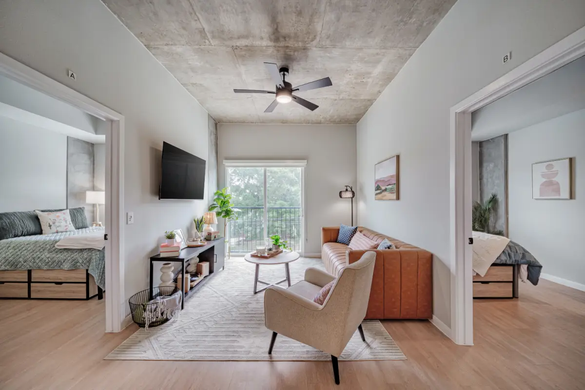 Modern living room with tan couch, armchair, and TV on the wall. Large window offers natural light. Bedrooms visible on either side, creating an inviting ambiance.