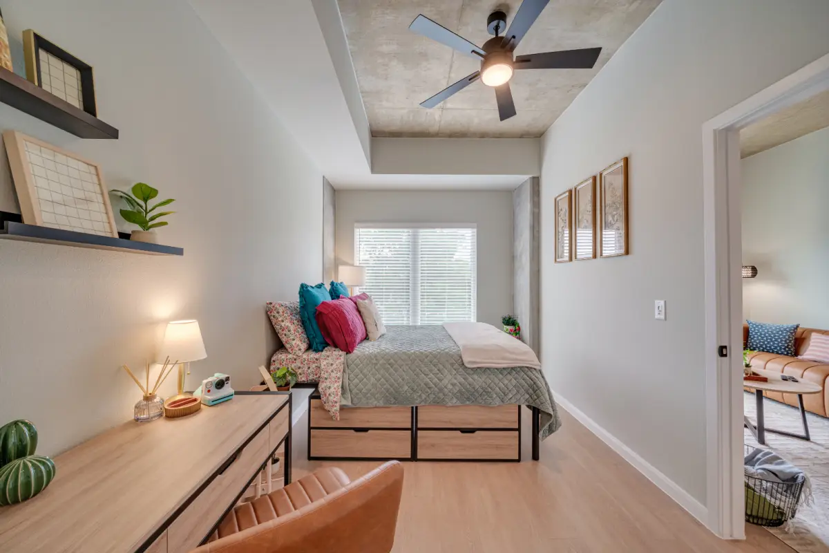 Modern bedroom with a large bed adorned with colorful pillows, a wooden desk with a lamp, wall shelves, and a ceiling fan. Bright and inviting atmosphere.