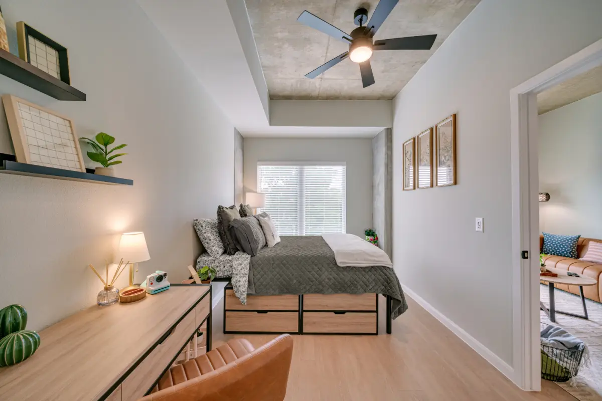 Modern bedroom with a large bed adorned with colorful pillows, a wooden desk with a lamp, wall shelves, and a ceiling fan. Bright and inviting atmosphere.