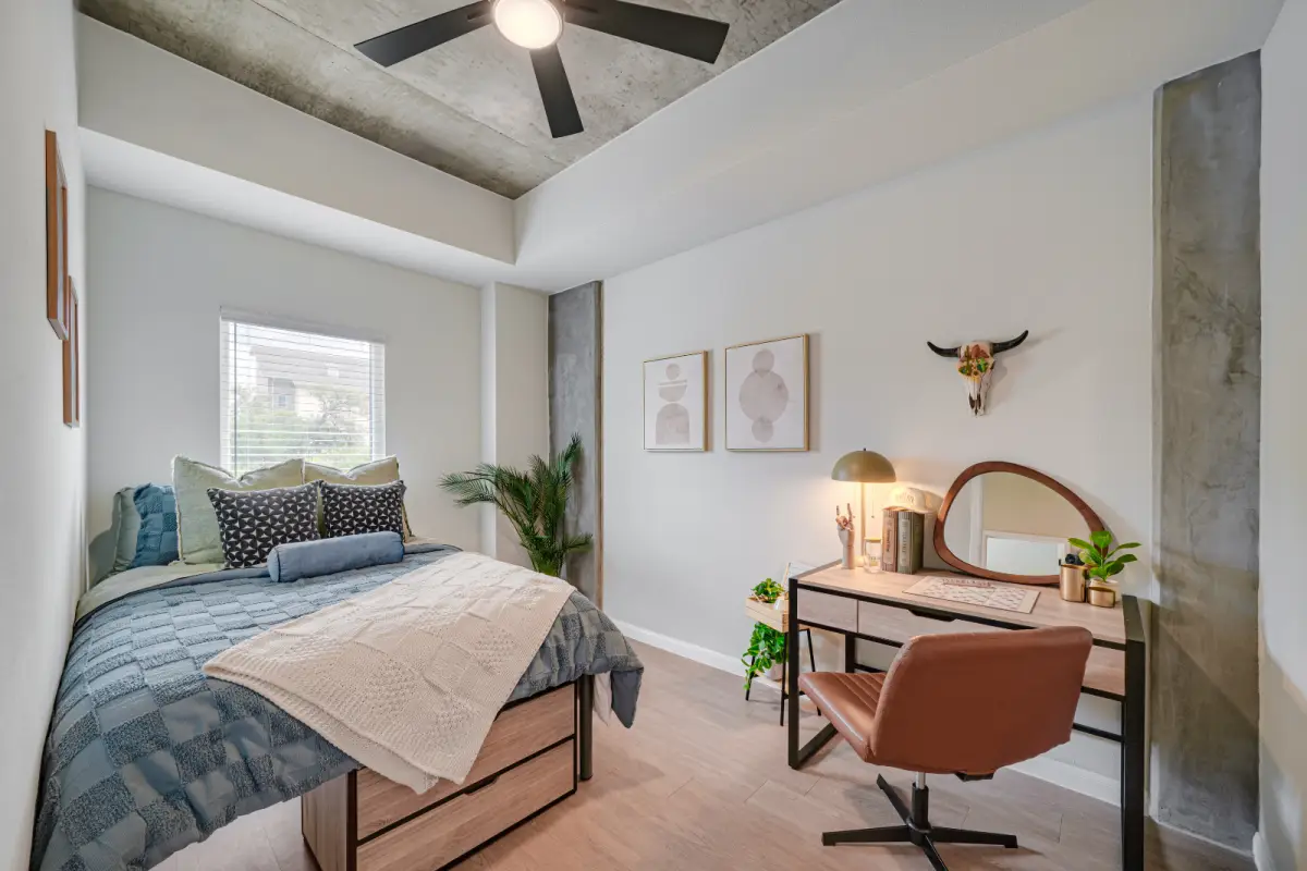 Modern bedroom with a large bed adorned with colorful pillows, a wooden desk with a lamp, wall shelves, and a ceiling fan. Bright and inviting atmosphere.