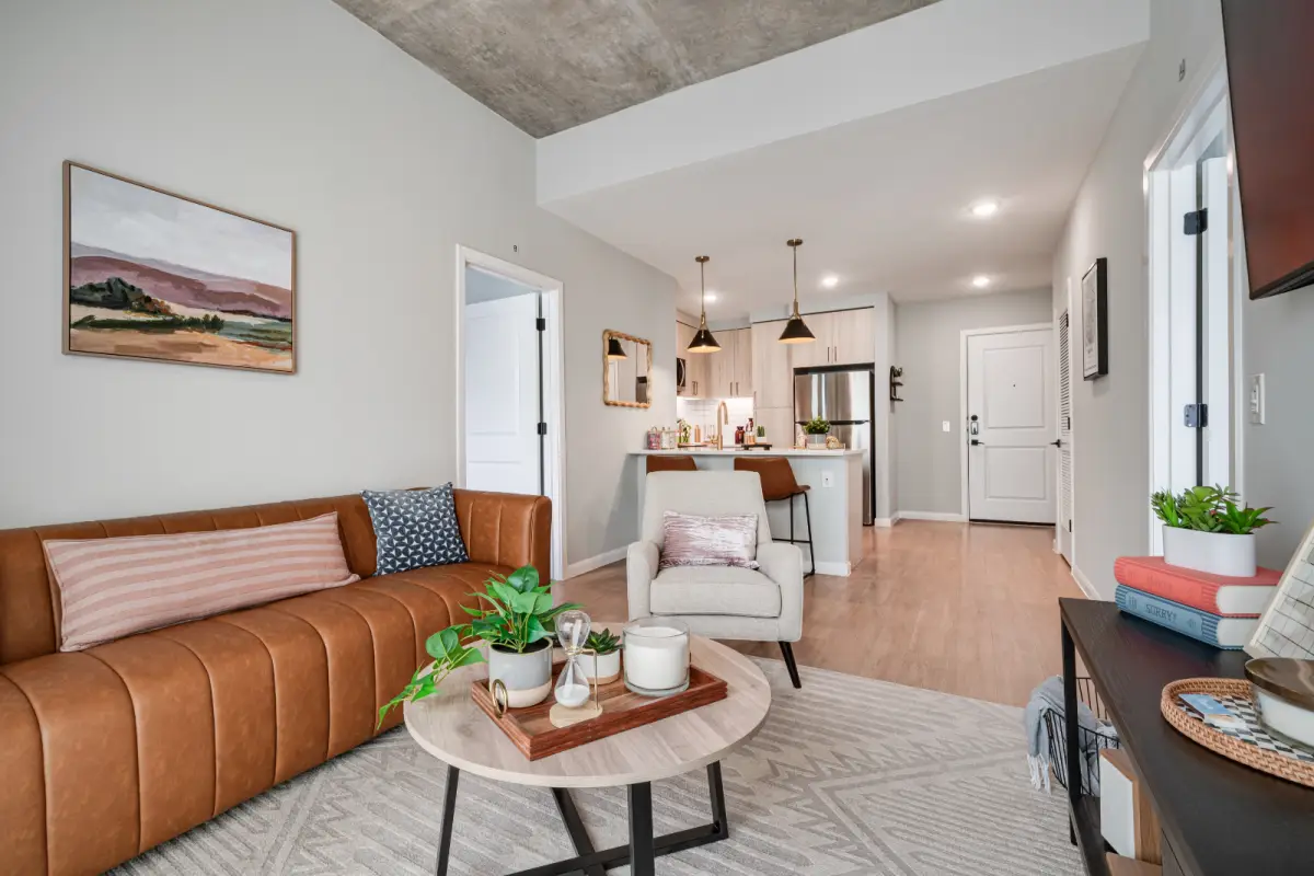 Modern living room with a brown leather sofa, patterned cushions, a round coffee table with candles and plants. Cozy kitchen in the background, neutral decor.
