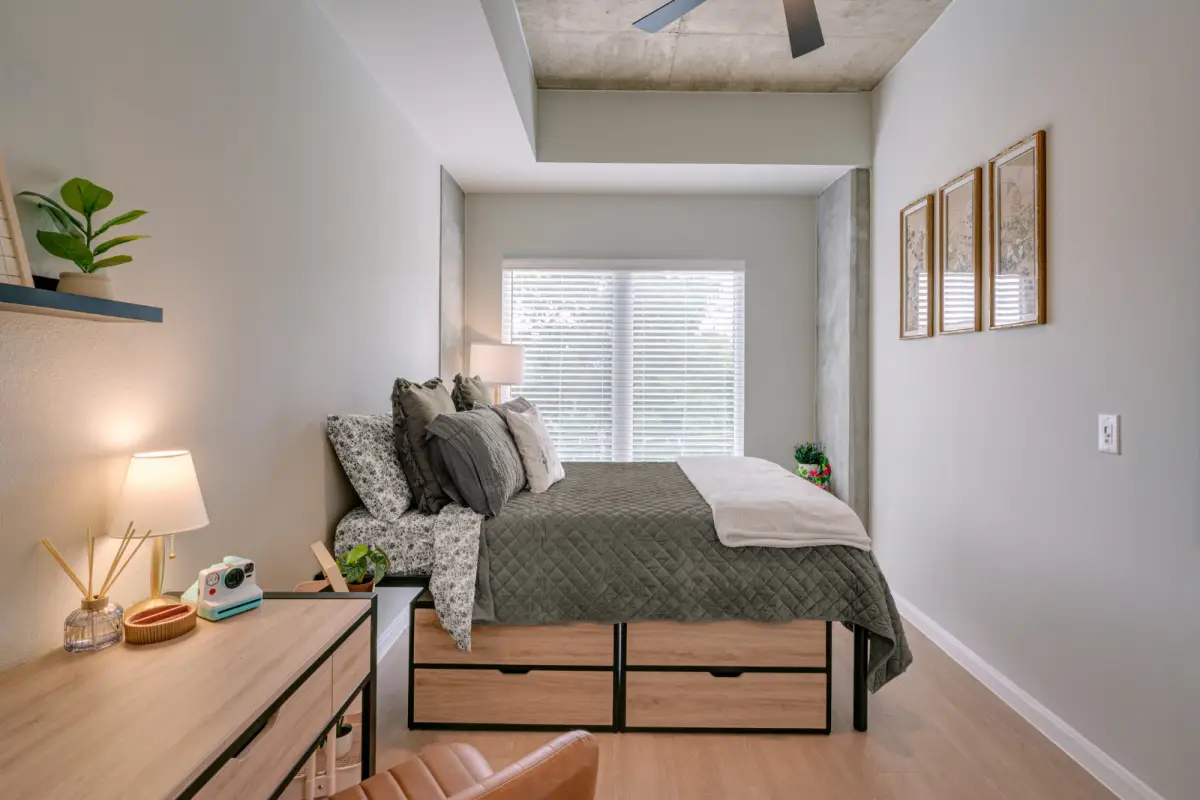Modern bedroom with a large bed adorned with colorful pillows, a wooden desk with a lamp, wall shelves, and a ceiling fan. Bright and inviting atmosphere.